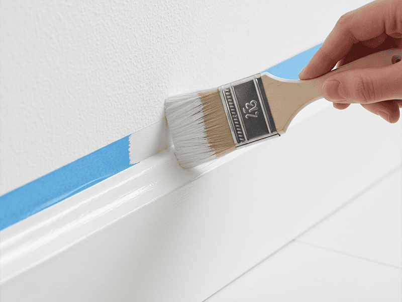An angled paintbrush applying white paint to a skirting board with a clean, taped edge.