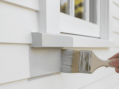 A paintbrush applying a coat of white paint to classic weatherboard siding.