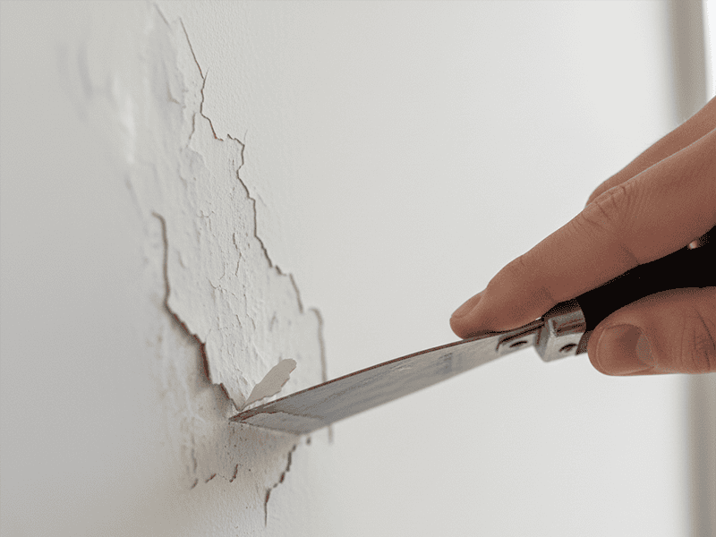 A hand using a metal scraper to carefully remove a section of bubbling paint from a wall.